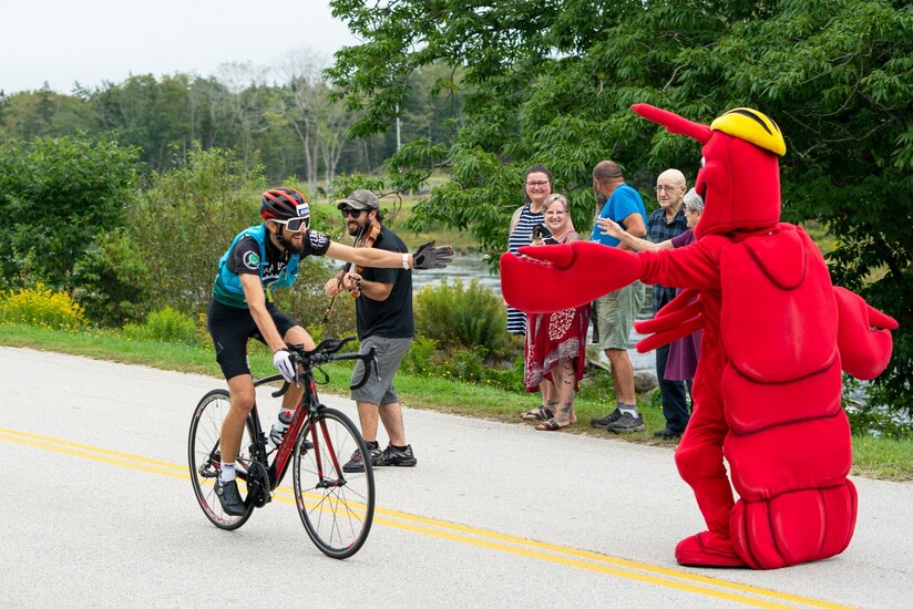 A picture of a cyclist high-fiving a lobster mascot, with people cheering on the cyclist and a violinist behind the cyclist.